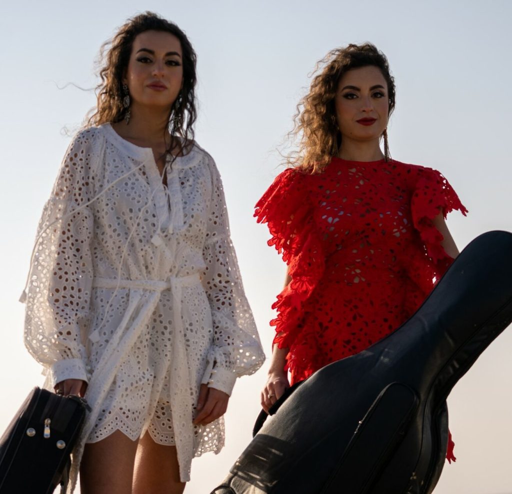 Two women standing next to each other both holding a cello, one on the left in a white dress and the one on the right in a red dress.