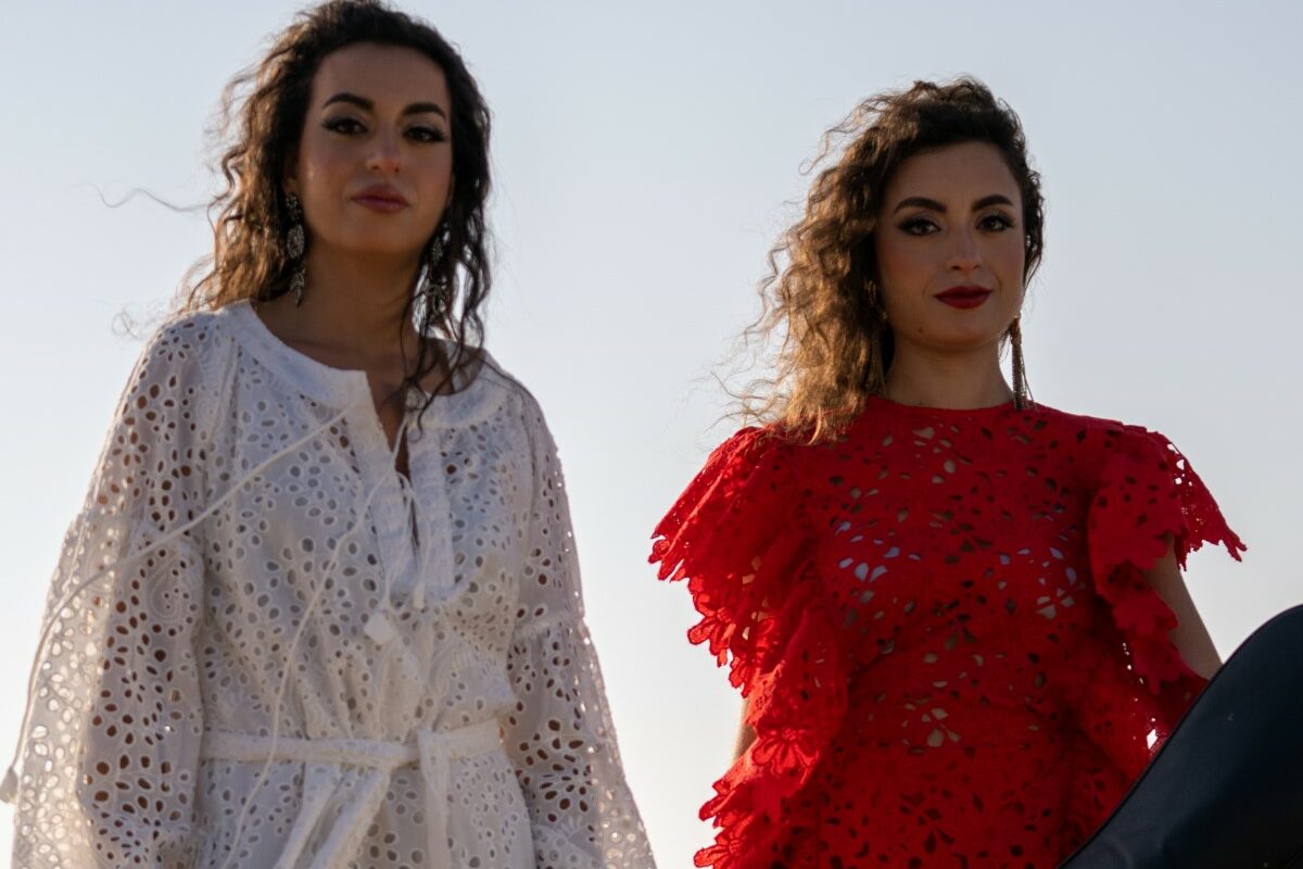 Two women standing next to each other both holding a cello, one on the left in a white dress and the one on the right in a red dress.
